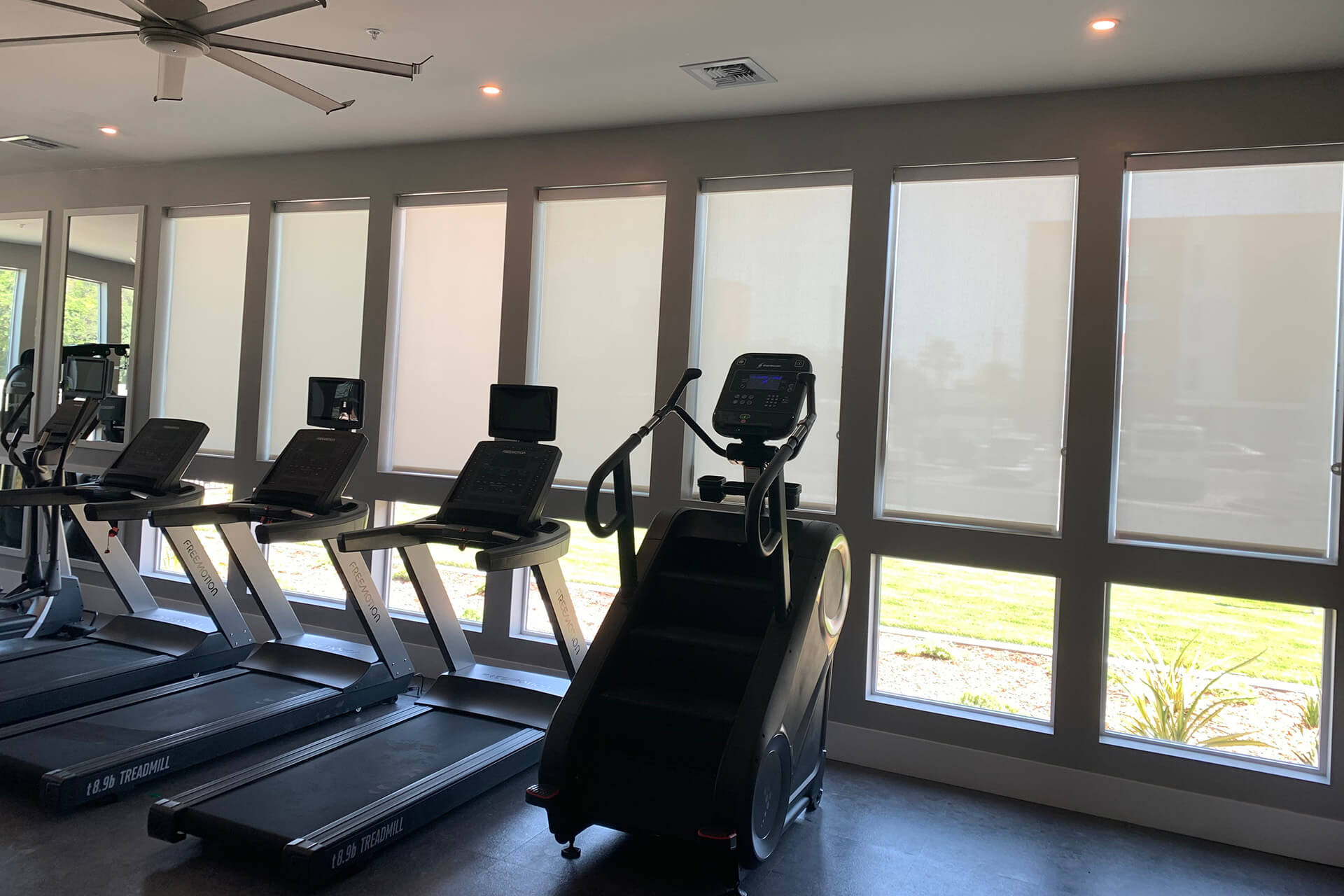 A gym with workout machines facing windows covered with roller shades.