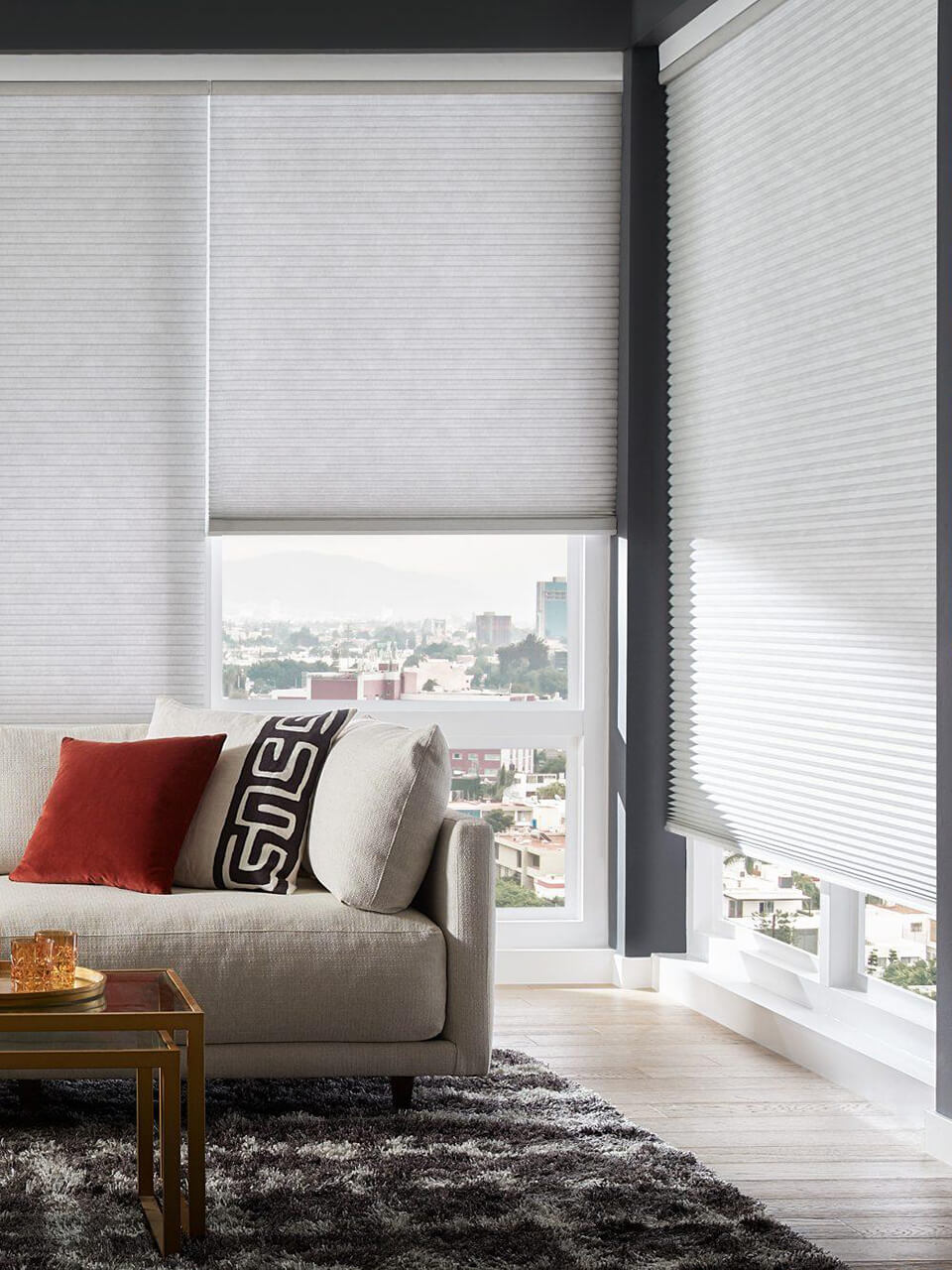The corner of a living room with white honeycomb shades half-covering the tall windows.