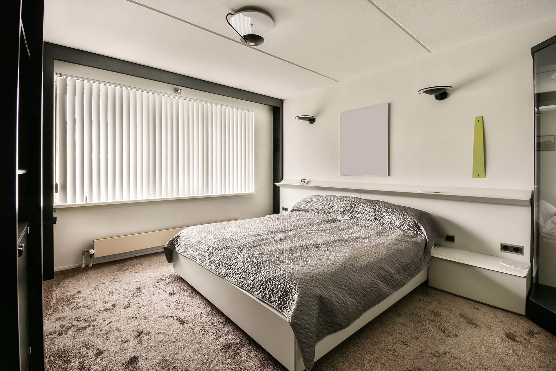 A minimalistic white bedroom with vertical blinds on the long window.